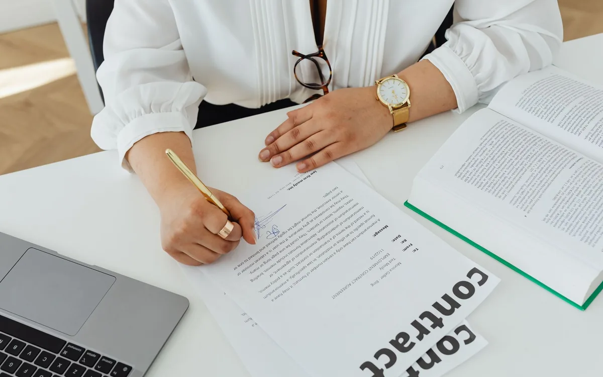 Business professional reviewing and signing a written contract beside a laptop